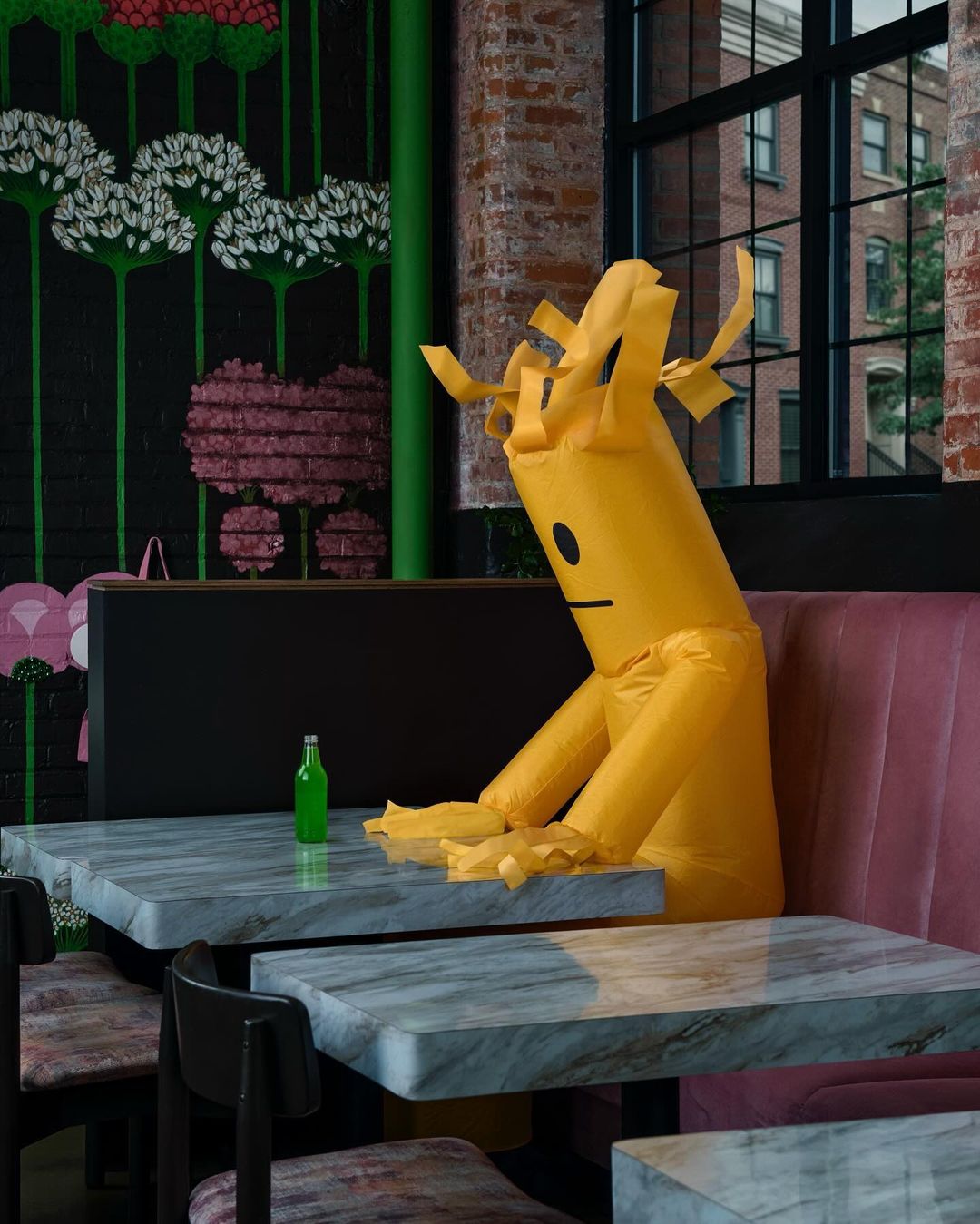 A yellow inflatable tube man sits alone at a marble table in a restaurant, looking forlorn with a bottle of soda.